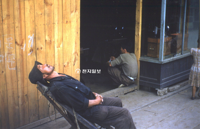 1954년 미국 종군기자가 찍은 사진으로 경기 수원지역 미싱가게 앞에 지게꾼이 일거리가 없자 지게에 기댄 채 절망스러운 표정으로 낮잠을 자고 있다. 그 옆에는 한 남성이 어깨가 축 쳐진듯 앉아 있다. (제공: 정성길 기록사진연구가) ⓒ천지일보 2022.2.25
